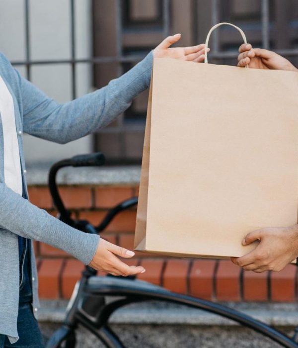 Food delivery, lunch to client and fast service. Millennial male courier sits on bicycle in company t-shirt, giving order to woman client in eco paper bag with order near house, cropped, panorama
